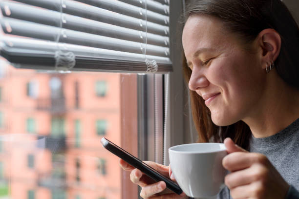 istock Happy woman with cup using phone near window 1349991192