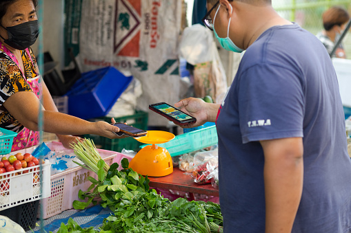 Thai man is paying contacless on local market in Bangkok. Vendor woman is reaching out mobile for scanning QR code. Both woman and man are wearing face masks.