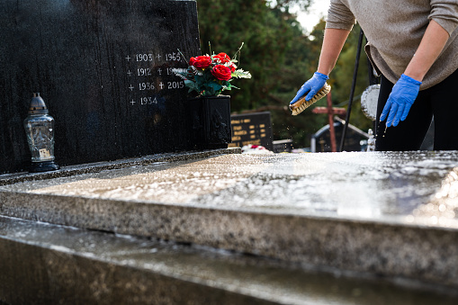 Unrecognizable woman washing the tombstone with a brush. Graveyard preparation for All Saints Day.