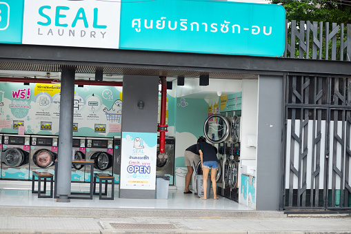 Young thai couple captured in open laundromat in Bangkok Chatuchak. View over street. People are filling washing machine. Laundromat is below condominum