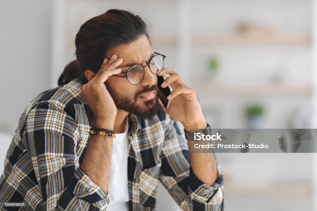 Upset middle-eastern guy having phone conversation, home interior Upset middle-eastern guy in casual sitting on couch, having phone conversation with lover or business partner, experiencing difficulties, home interior, copy space, closeup portrait Using Phone Stock Photo
