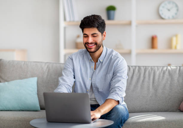 istock Home office concept. Happy arab guy working on pc laptop, sitting on comfy sofa at home interior, free space 1350011502
