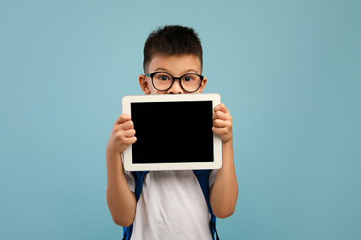 Distant Education. Asian Schoolboy Holding Digital Tablet With Blank Black Screen, Recommending New Educational App Or Website While Posing Over Blue Studio Background, Mockup Image With Copy Space