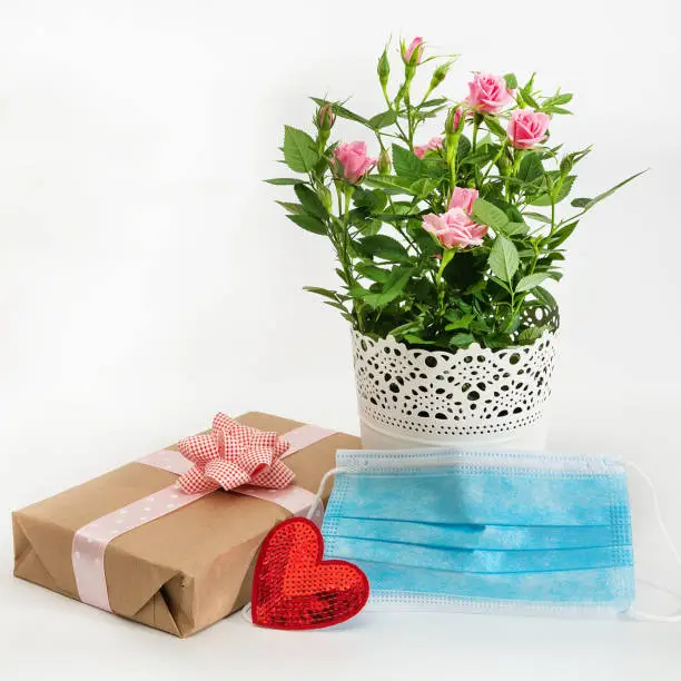 Photo of A bouquet of roses, a medical mask, a valentine card and a gift on a white background. Holidays concept during coronavirus period