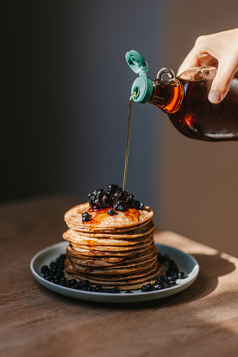 Hand pouring maple syrup on oatmeal pancakes topped with wild blueberries