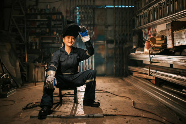 Asian chinese smiling female welder with protective workwear  garage sitting on stool in the workshop stock photo