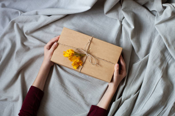 Hands hold beautiful gift box on a bed. Simple gift packaging, craft box, ribbon twine, dry yellow flower, on grey linen fabric background. stock photo