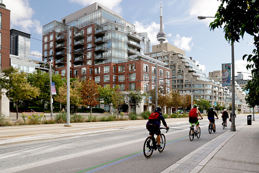 Toronto, Canada - September 3, 2021: Cyclists head east on the bicycle lane along the 500-block Queens Quay West in Harbourfront. Very little traffic during the fourth wave of COVID-19.\n\nUpscale residential buildings line the Waterfront Communities. Background shows the CN Tower, also illustrated on a colourful street banner. Summer morning with light cloud cover in the downtown district.