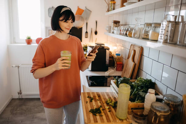 istock Pretty young woman using her mobile phone while drinking detox juice in the kitchen at home 1350041647