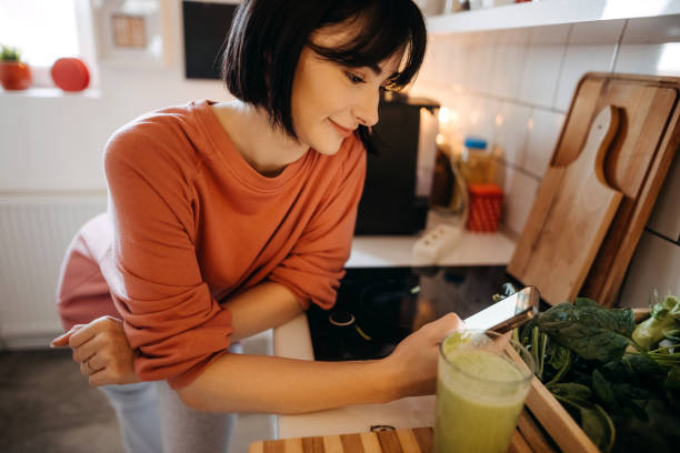 istock Pretty young woman using her mobile phone while drinking detox juice in the kitchen at home 1350041651
