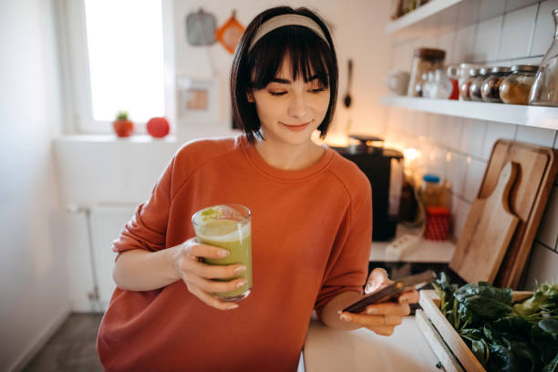 istock Pretty young woman using her mobile phone while drinking detox juice in the kitchen at home 1350041664