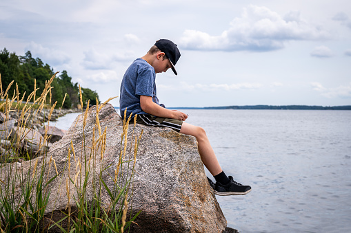 Side view of young boy sitting on rock at seashore looking at mobile phone. Water and horizon in the background. Tranquil scene in Sweden.