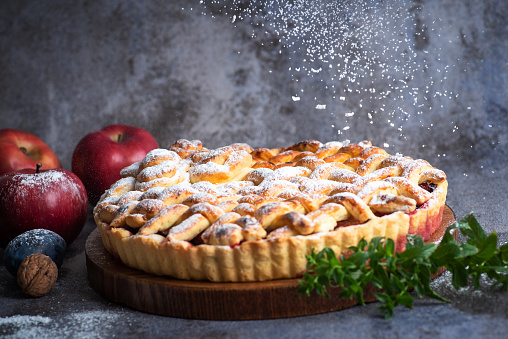 Autumn apple pie with decorated crust surrounded with leaves and red apples on wooden table.
