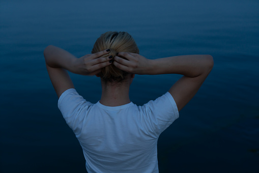 The blonde girl looks at the sea after sunset and straightens her hair