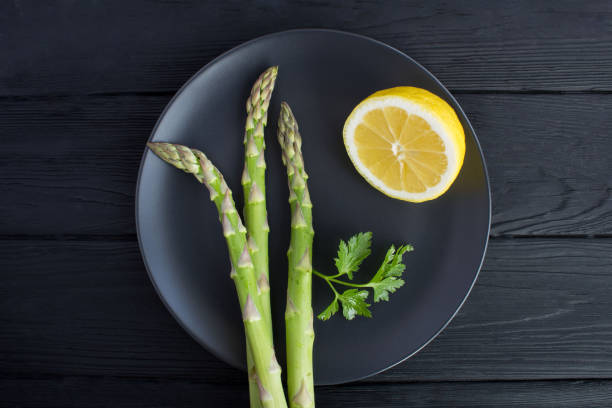 Raw asparagus and lemon in the dark plate on the black wooden background. Top view.