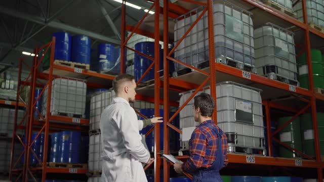 Rear view tracking shot of engineer in white coat explaining task to young warehouse worker standing by steel racks with containers and barrels in industrial storage