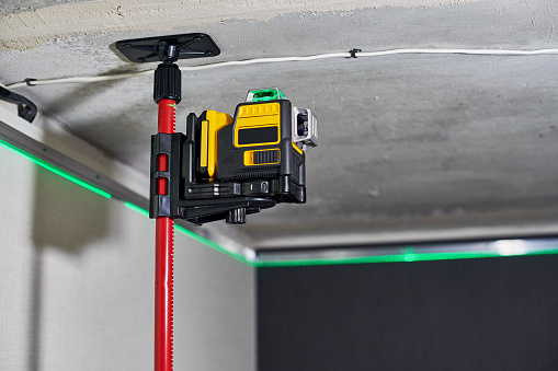 Laser level measuring tool with visible green laser beam on wall at unfinished apartment. Professional equipment for installing stretch ceiling. Home renovation concept, close up