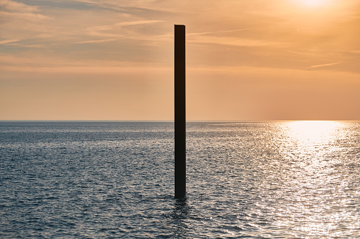 Large rusty steel construction pillar in blue sea with calm waves at sunset, sunlight reflecting on seawater surface. Vertical abandoned building part, construction site near coast