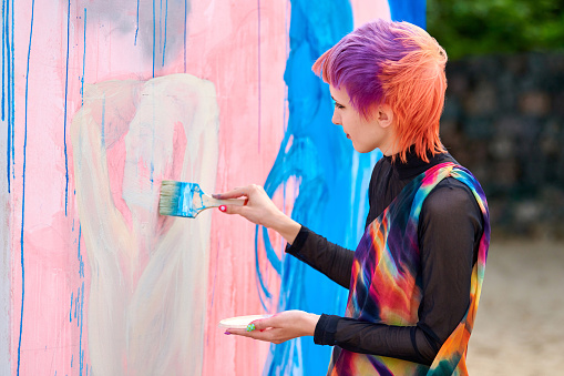 Svetlogorsk, Russia - 08.14.2021 - Woman artist with short multicolored hair in bright colorful clothes painting beautiful picture with paint brush. Oil painting on large canvas with blue, pink colors