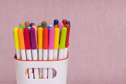 multicolored markers in a white glass on a pink background, copy space. School children's drawing supplies