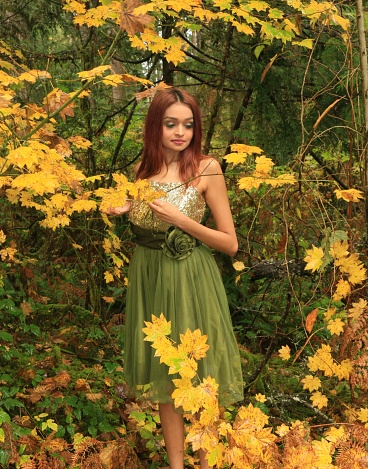 A woman posing in yellow Autumn leaves wearing a green and gold sleeveless dress.