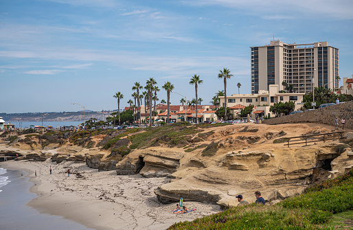 La Jolla, California, USA - October 3, 2021: brown stone rock formation and caves on Wipeout Beach with tall condominium building above under blue cloudscape.