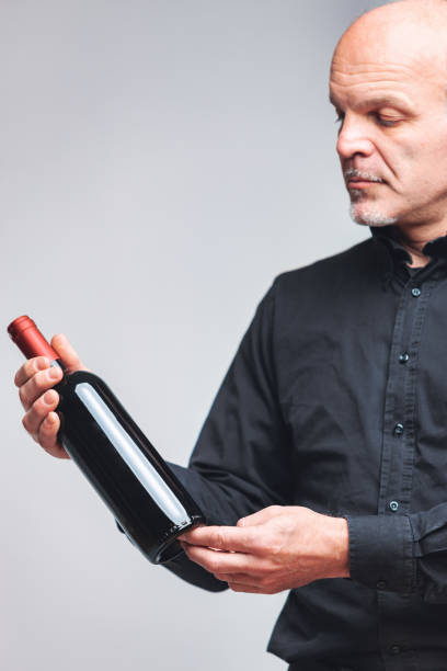 Thoughtful senior man eyeing up a bottle of red wine Thoughtful senior man eyeing up an unlabelled bottle of red wine held in his hands with a pensive expression over grey in a close up cropped portrait taste test stock pictures, royalty-free photos & images