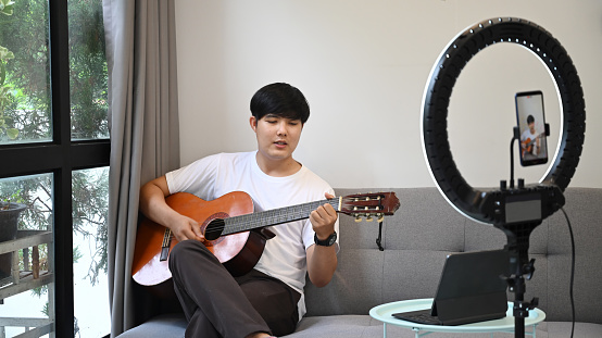 Young asian man playing guitar and streaming online vlog at home.