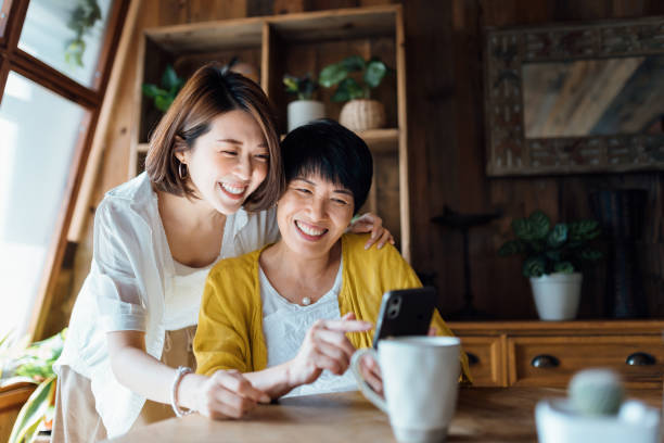 affettuosa madre e figlia asiatica che usano lo smartphone insieme a casa, sorridono gioiosamente, si godono il tempo di legame tra madre e figlia. famiglia e tecnologia multigenera generazione - smart phone asian ethnicity mobile phone women foto e immagini stock