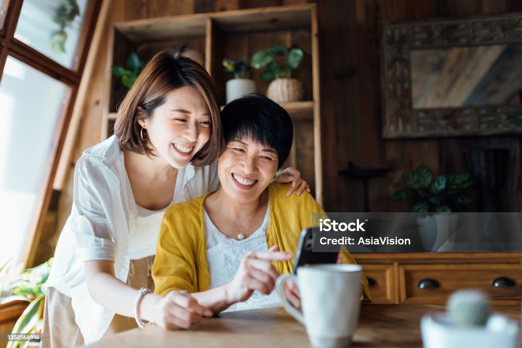 Affectionate Asian senior mother and daughter using smartphone together at home, smiling joyfully, enjoying mother and daughter bonding time. Multi-generation family and technology Asian and Indian Ethnicities Stock Photo