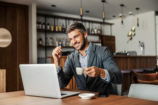 A happy businessman is sitting in a cafe, drinking coffee and watching a webinar on the laptop. The man has earphones in his ears so he can listen to the lecture. A businessman using laptop in cafe