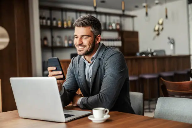 Photo of A happy businessman is sitting in a coffee shop and checking on his bank account on the mobile. There is a laptop on a table. A man using the phone for e-banking