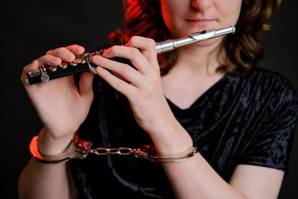 istock Woman musician with a flute in handcuffed hands on a studio dark background. Problems of musical playing at concerts, the concept of non-freedom and arrests 1350178100