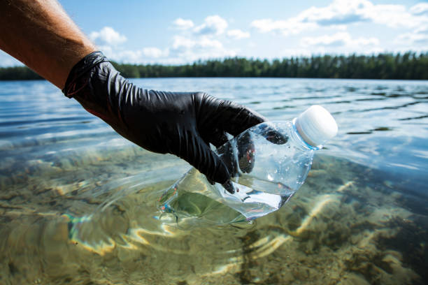 istock Hand in rubber glove picking up used plastic bottle floating in clear water. 1350185532