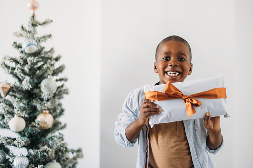 Happy smiling African American boy standing next to the Christmas tree and holding wrapped gift in hands while looking at camera