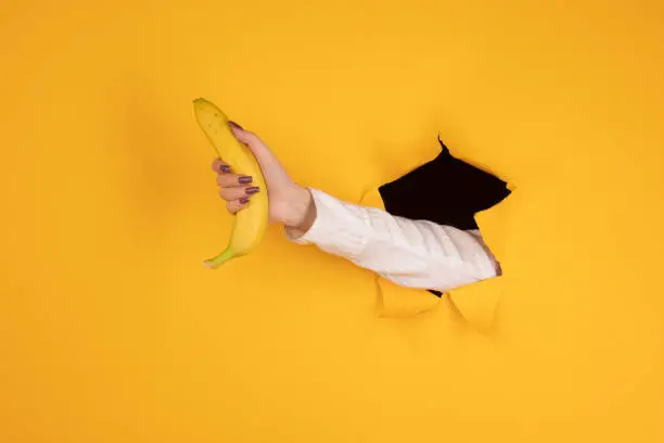 A woman holds a banana in her hand, inserted through a hole in torn yellow paper