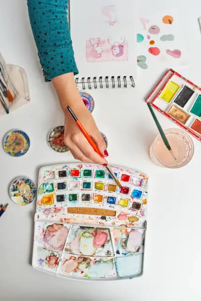 Top view of unrecognizable woman's hands making illustrations with watercolors working from home in her office