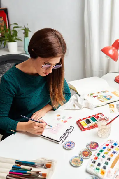 Woman painting an illustration with watercolors working from home sitting at the desk