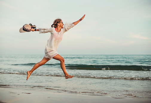 a middle-aged woman jumps on the seashore. she is in profile with her legs and arms outstretched. she is wearing a white dress and hat in her hand. she is happy and contented.
