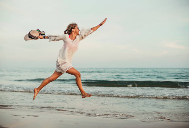 a middle-aged woman jumps on the seashore. she is in profile with her legs and arms outstretched. she is wearing a white dress and hat in her hand. she is happy and contented. - water human hand people women imagens e fotografias de stock