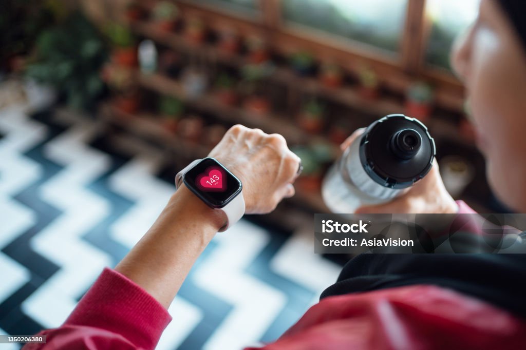 Over the shoulder view of senior Asian woman resting after exercising at home, looking at her smart watch, using fitness tracker app and measuring pulse. Maintaining healthy fitness habits. Elderly wellbeing, health, wellness and technology concept Wellbeing Stock Photo