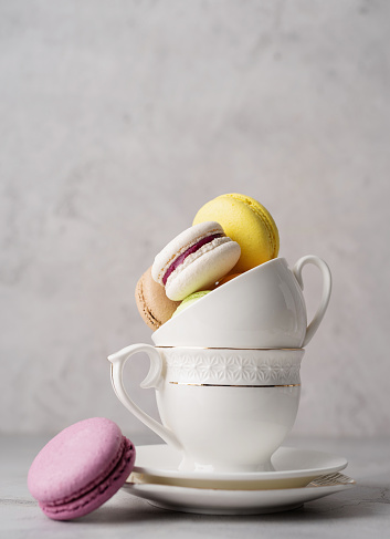 Stack of white coffee cups filled with French dessert colorful macaroons on the edge of the table, white brick wall background