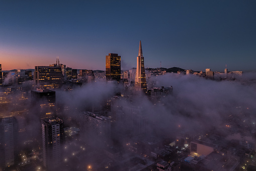 Dramatic aerial view above the fog in San Francisco's financial district. Skyscrapers rise up through the heavy fog and lights twinkle below as the sun comes up.