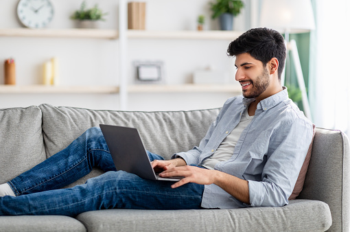 Happy arab guy resting with laptop on sofa at home interior, copy space. Handsome man using notebook, watching movie, surfing internet or scrolling social networks