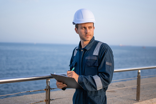 Serious young male engineer in hardhat and protective clothes looking away while inspecting sea port and taking notes on clipboard