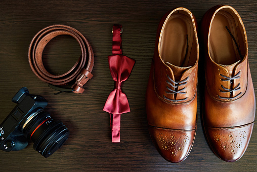 Close up of brown leather male shoes, bow tie, belt and camera on wooden background. Modern man or groom accessories. Wedding details. Male casual outfits. Top view, flat lay