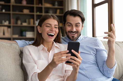 Overjoyed young family couple looking at telephone screen, feeling excited getting shopping discount sale deal offer, celebrating reading online lottery gambling win notification, sitting on sofa.