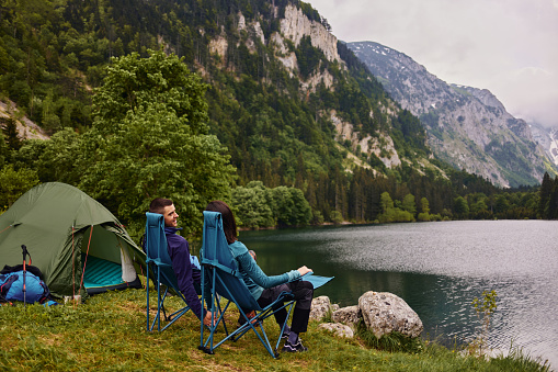 Relaxed couple sitting in camping chairs outside tent while camping by the lake