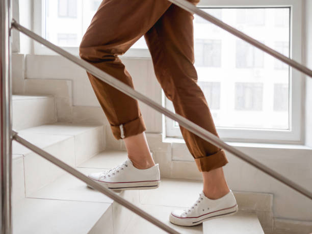 Woman in white sneakers and khaki trousers goes upstairs to her apartment. White staircase in apartment building. Casual outfit, urban fashion. stock photo