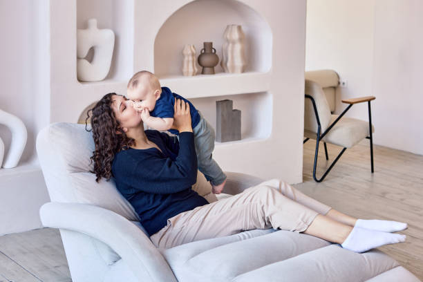 Woman spends time with infant on sofa in living room. Young mother holds little boy while sitting on couch in lounge during maternity leave. chaise longue photos stock pictures, royalty-free photos & images